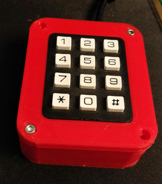 Photo of the old macropad. It's a particularly low quality telephone keypad in a very tall red 3d printed case with lots of visible sharp edges. There are places for four screws at the corners but only two have been installed.