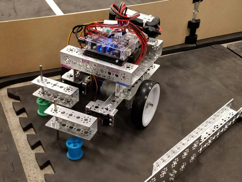 Photo of a small robot made of aluminum pieces stuck together with screws, sitting on a foam tile. There is a wall used in the course visible in the background. There is indistinct handwriting visible on the front of the robot. The robot has a controller circuit board on the top of it and two servo motors on the front with arms attached to them. Near the arms, there are brightly coloured spools as would be used for thread for sewing.